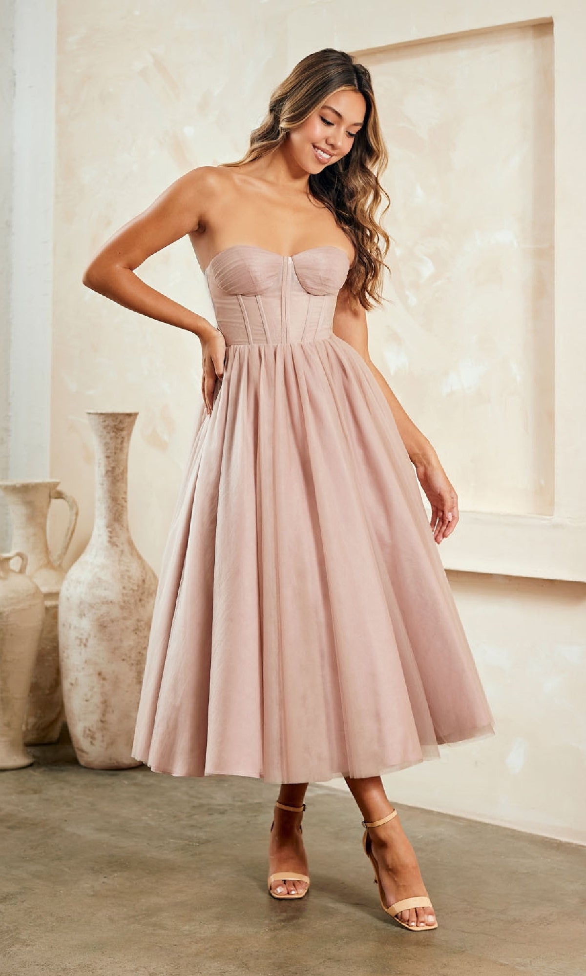 A woman stands indoors near large decorative vases, wearing the Strapless Pink Semi-Formal Dress: Amelia Couture, featuring a fitted bodice and flared tea-length skirt.